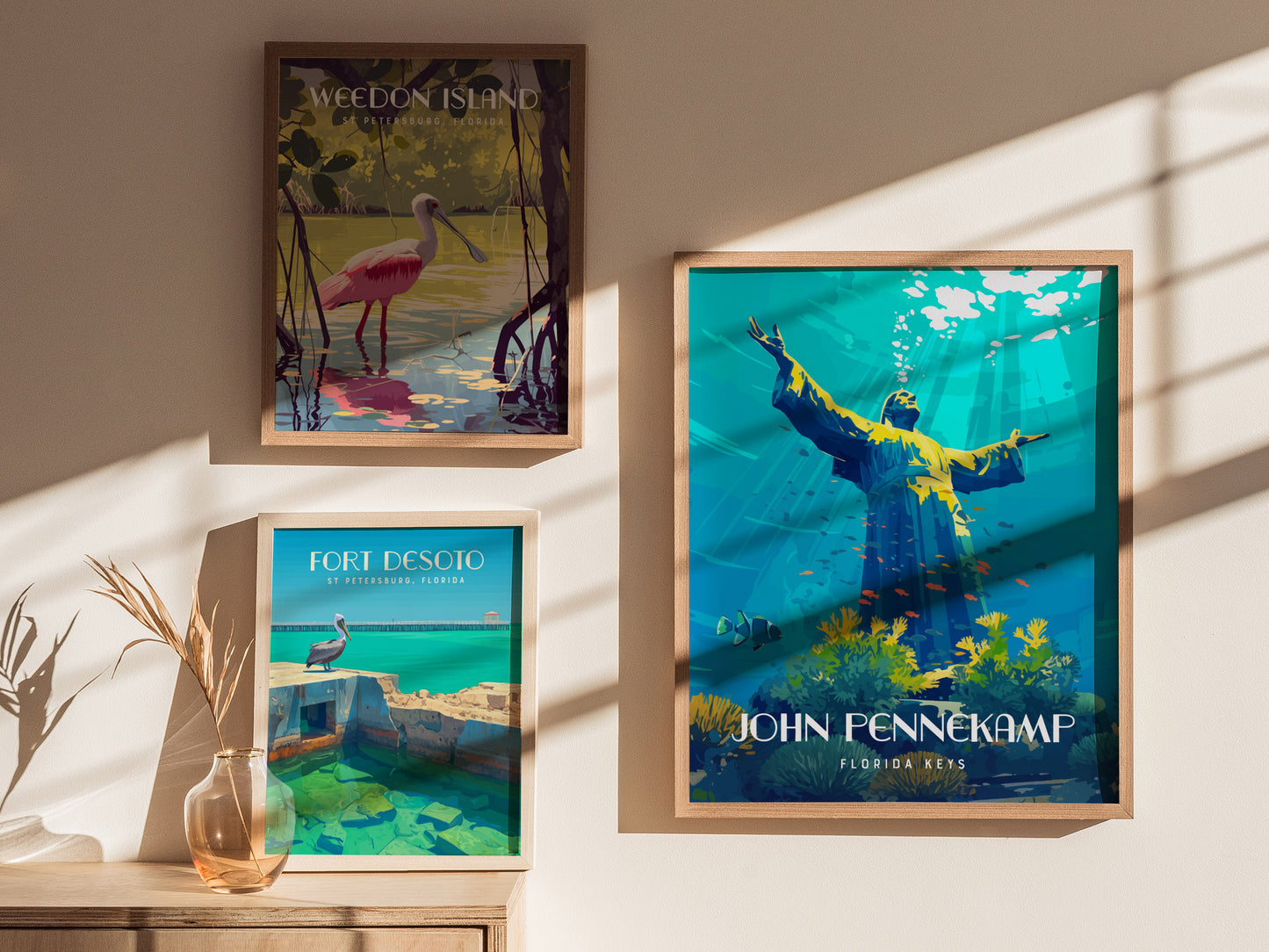 Three framed posters on a wall depict Weedon Island, Fort Desoto, and John Pennekamp Coral Reef State Park. Sunlight casts shadows, and a vase with dried plants sits below.