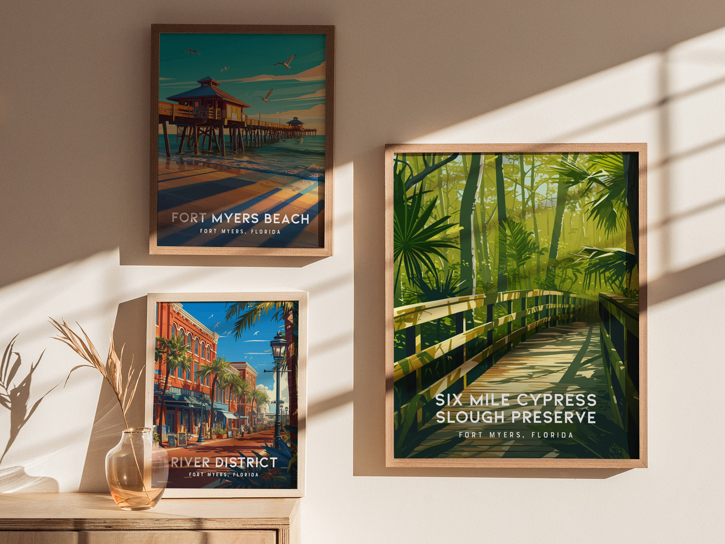 Three framed posters are displayed on a wall, featuring Fort Myers Beach, River District, and Six Mile Cypress Slough Preserve, with shadows and a decorative vase nearby.