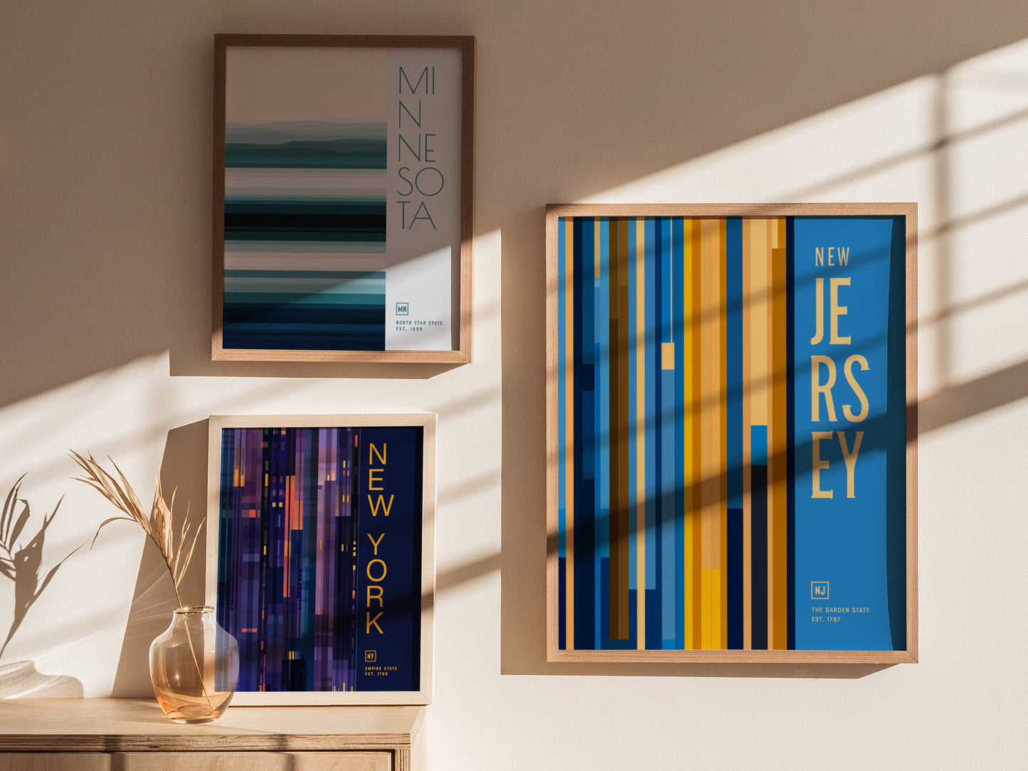 Framed posters of Minnesota, New York, and New Jersey hang on a sunlit wall, each featuring colorful vertical and horizontal lines.