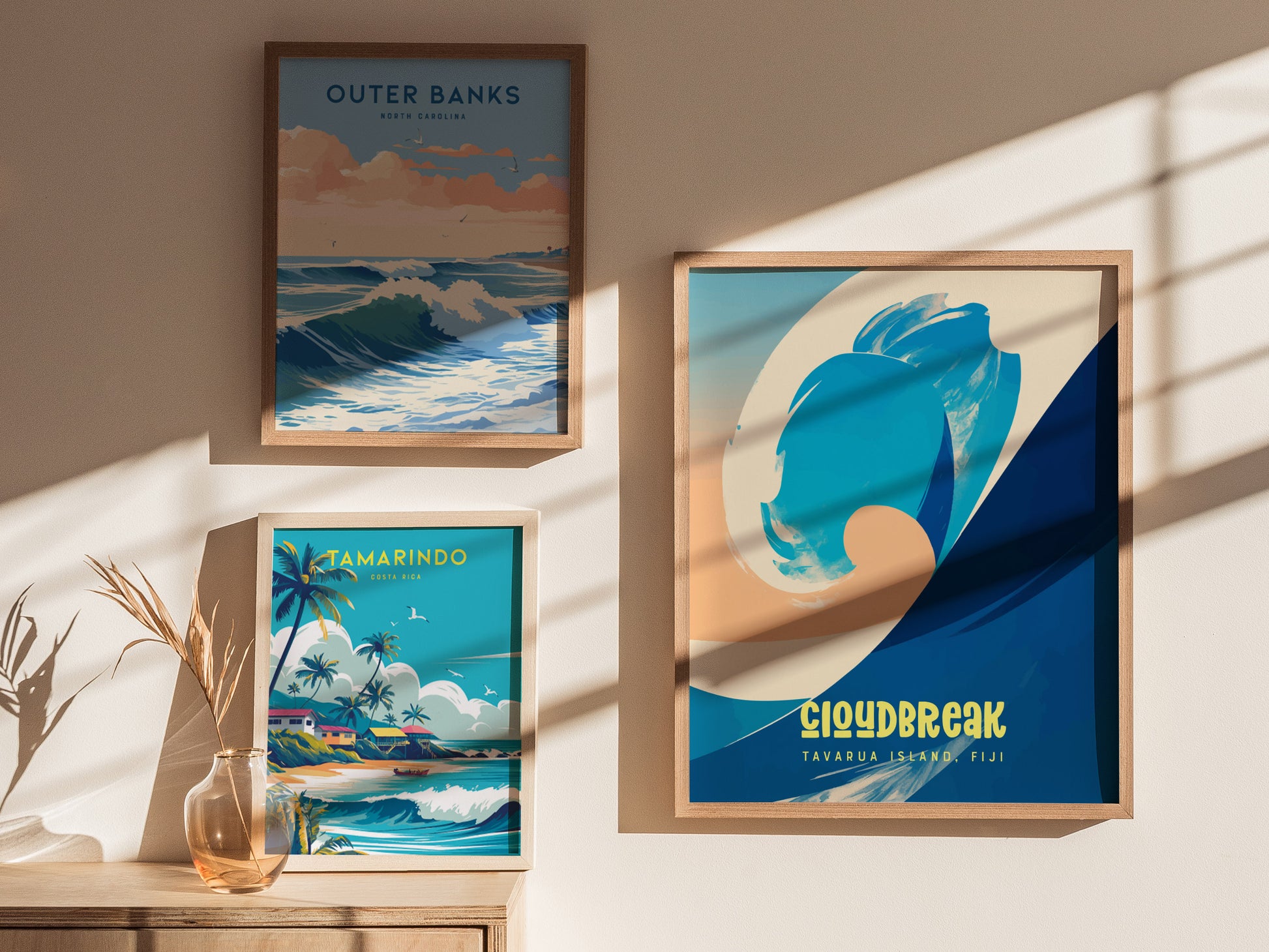 Three framed surf posters are displayed on a sunlit wall. The artworks depict scenes from Outer Banks, Tamarindo, and Cloudbreak with vibrant coastal imagery and text.