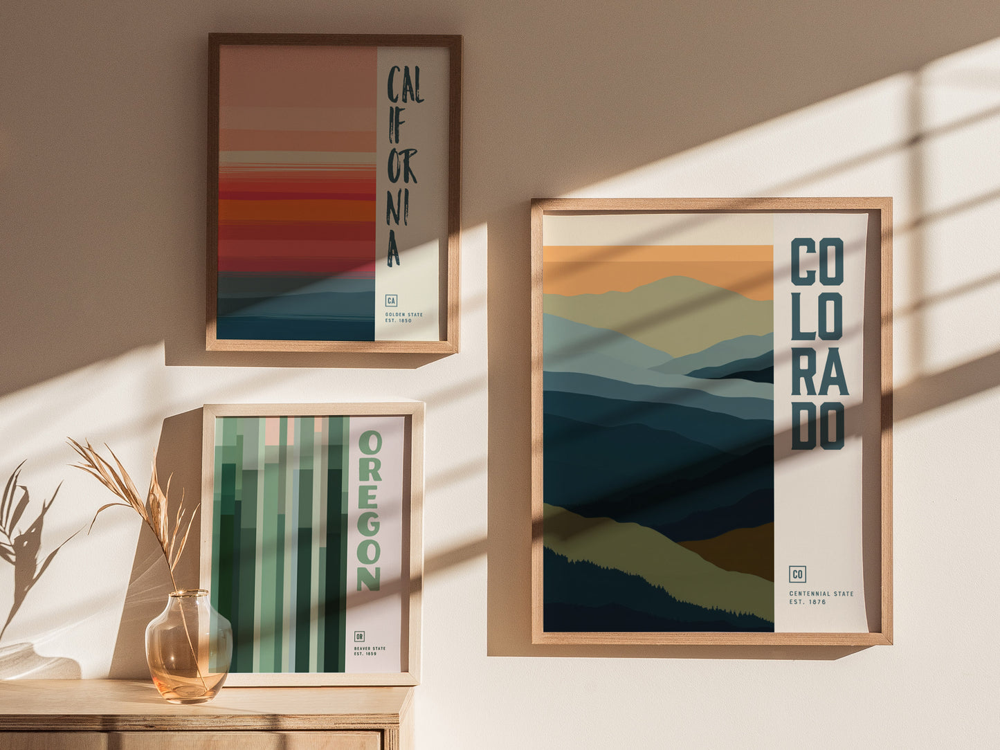 Three framed minimalist posters featuring abstract designs of California, Colorado, and Oregon hang on a sunlit wall. A small vase with dried grasses sits on a wooden table.