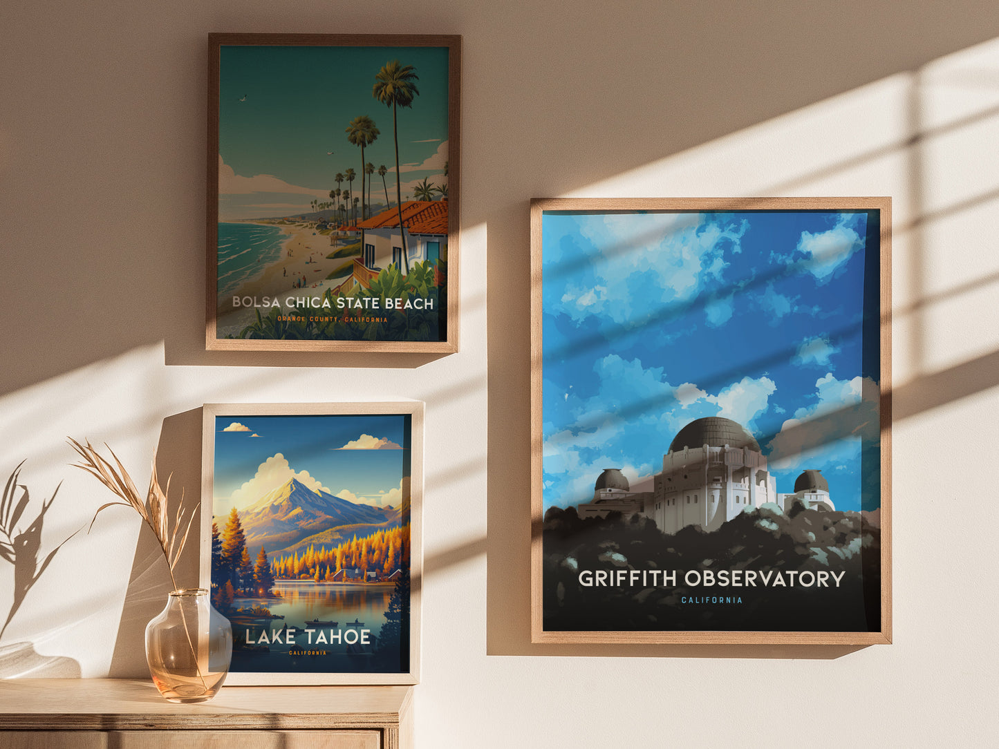 Three framed posters are displayed on a wall, featuring Bolsa Chica State Beach, Lake Tahoe, and Griffith Observatory, with natural light casting shadows across them.
