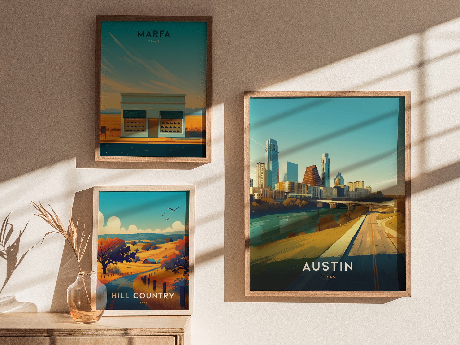 Three framed posters are displayed on a sunlit wall, featuring Marfa, Hill Country, and Austin, Texas landscapes. A glass vase with dried grass sits on a wooden surface.