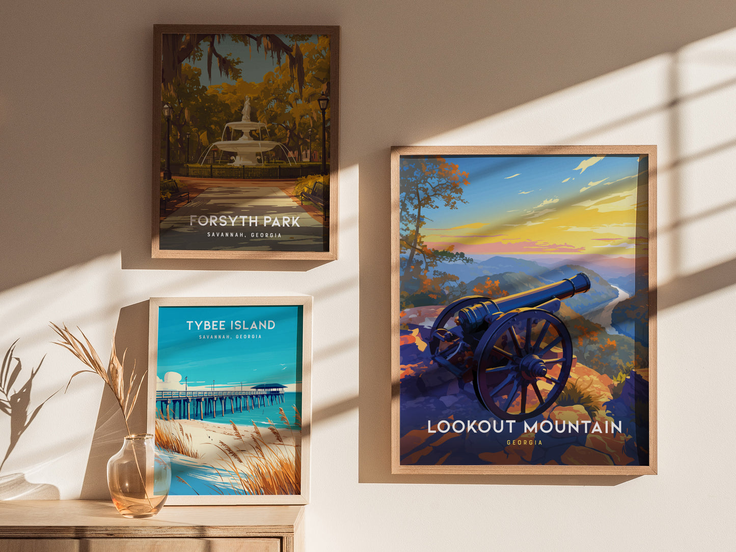 Framed posters of Forsyth Park, Tybee Island, and Lookout Mountain are displayed on a sunlit wall. A decorative vase with dried grass sits on a wooden surface below.