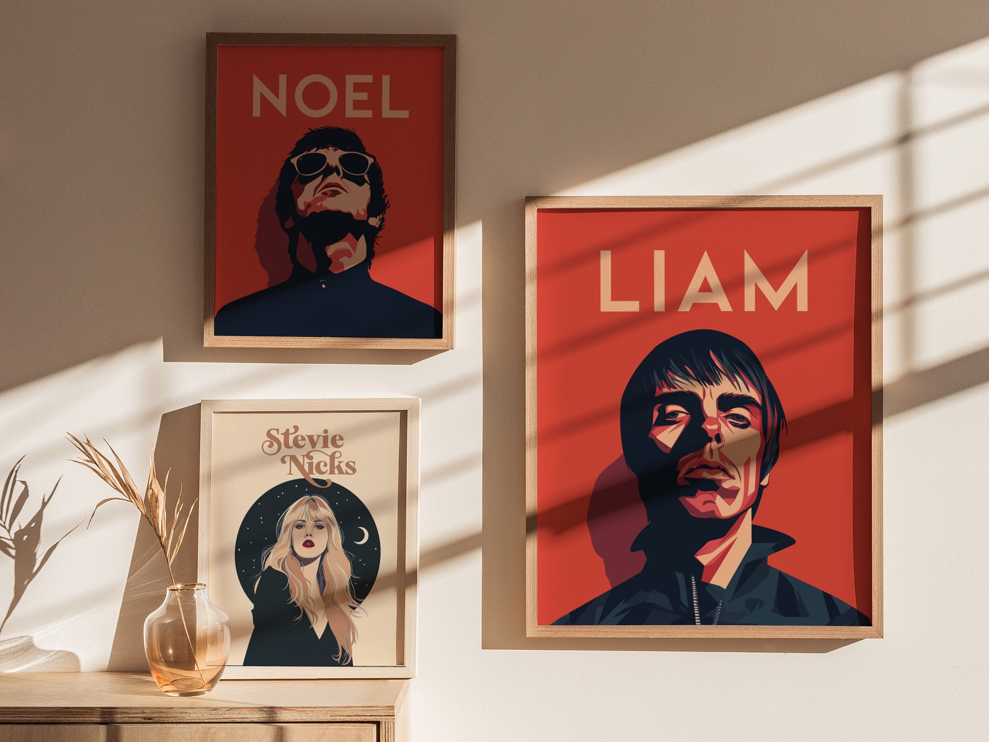 Three framed posters on a wall include stylized portraits labeled Noel, Liam, and Stevie Nicks. A glass vase with dried grass is on a wooden surface below.