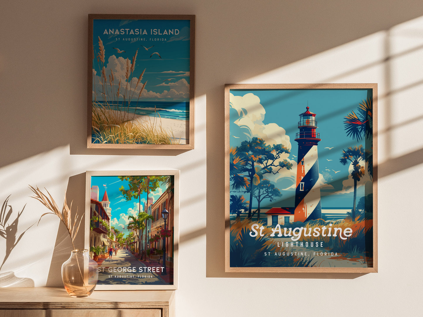 Three framed posters are displayed on a sunlit wall. The center poster features the St. Augustine Lighthouse with a spiral design, surrounded by palm trees. The other posters depict Anastasia Islands beach scene and a vibrant St.
