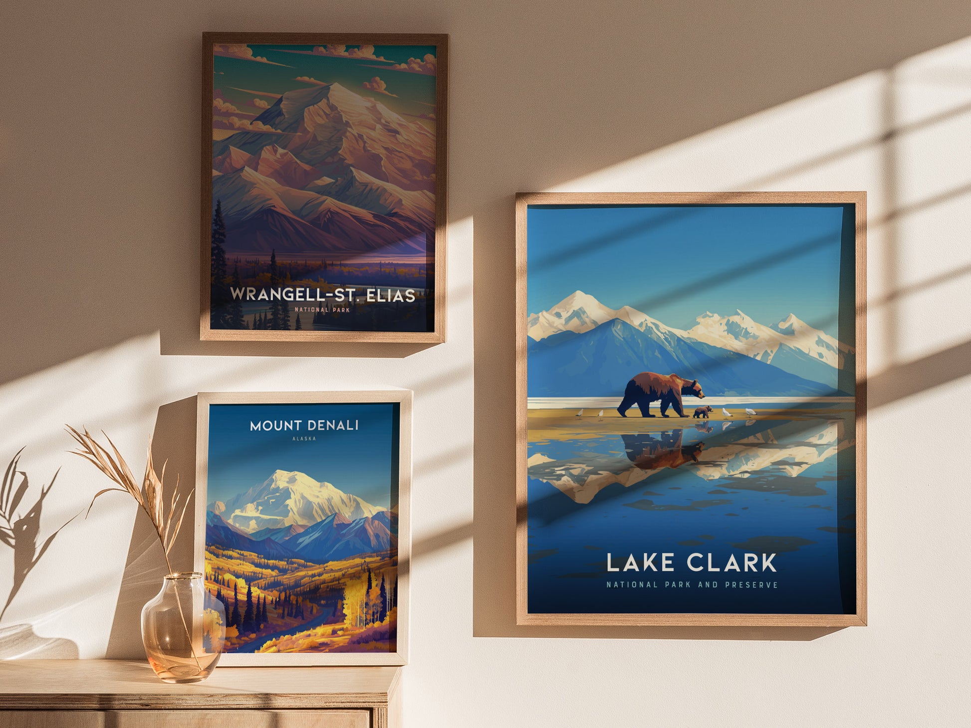 Three framed posters on a wall depict national parks: Wrangell-St. Elias, Mount Denali, and Lake Clark, featuring scenic mountains and wildlife art. Soft shadows and a vase add warmth.