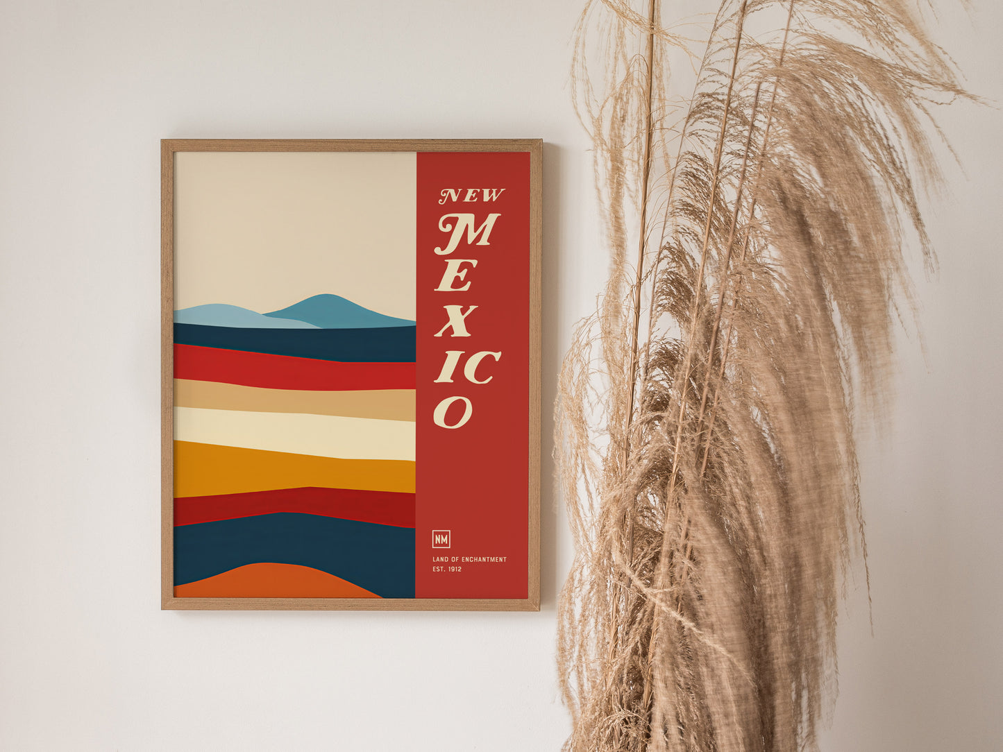 A framed poster with colorful horizontal stripes and the text New Mexico in bold letters, next to decorative pampas grass against a light wall.