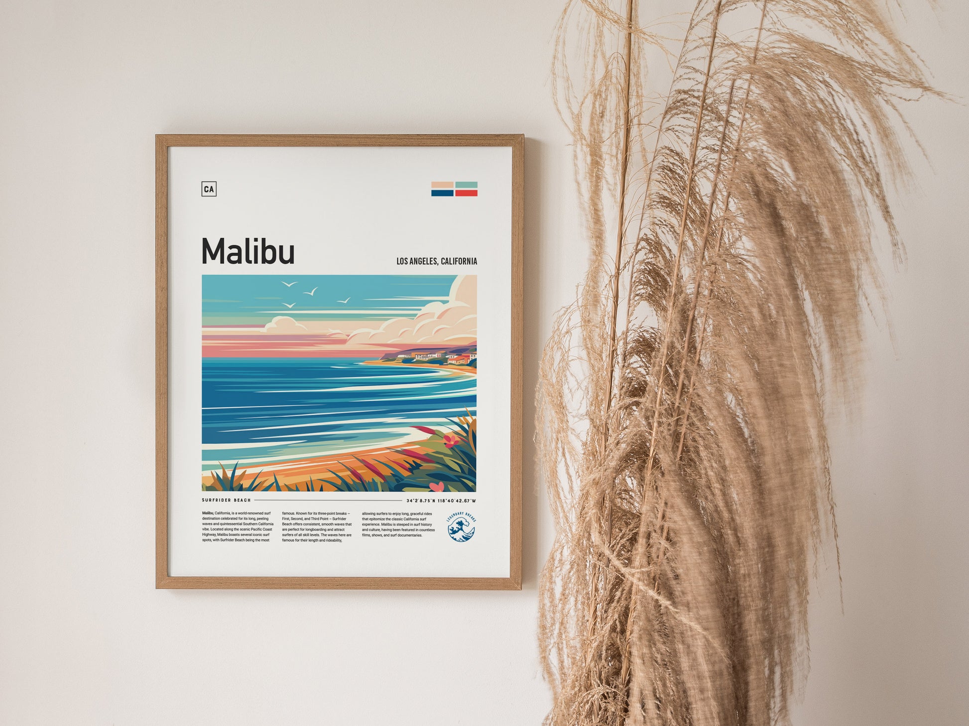 Framed poster of Malibu, California, featuring an abstract illustration of a coastline with sea, sky, and clouds, displayed beside tall dried grasses against a white wall.