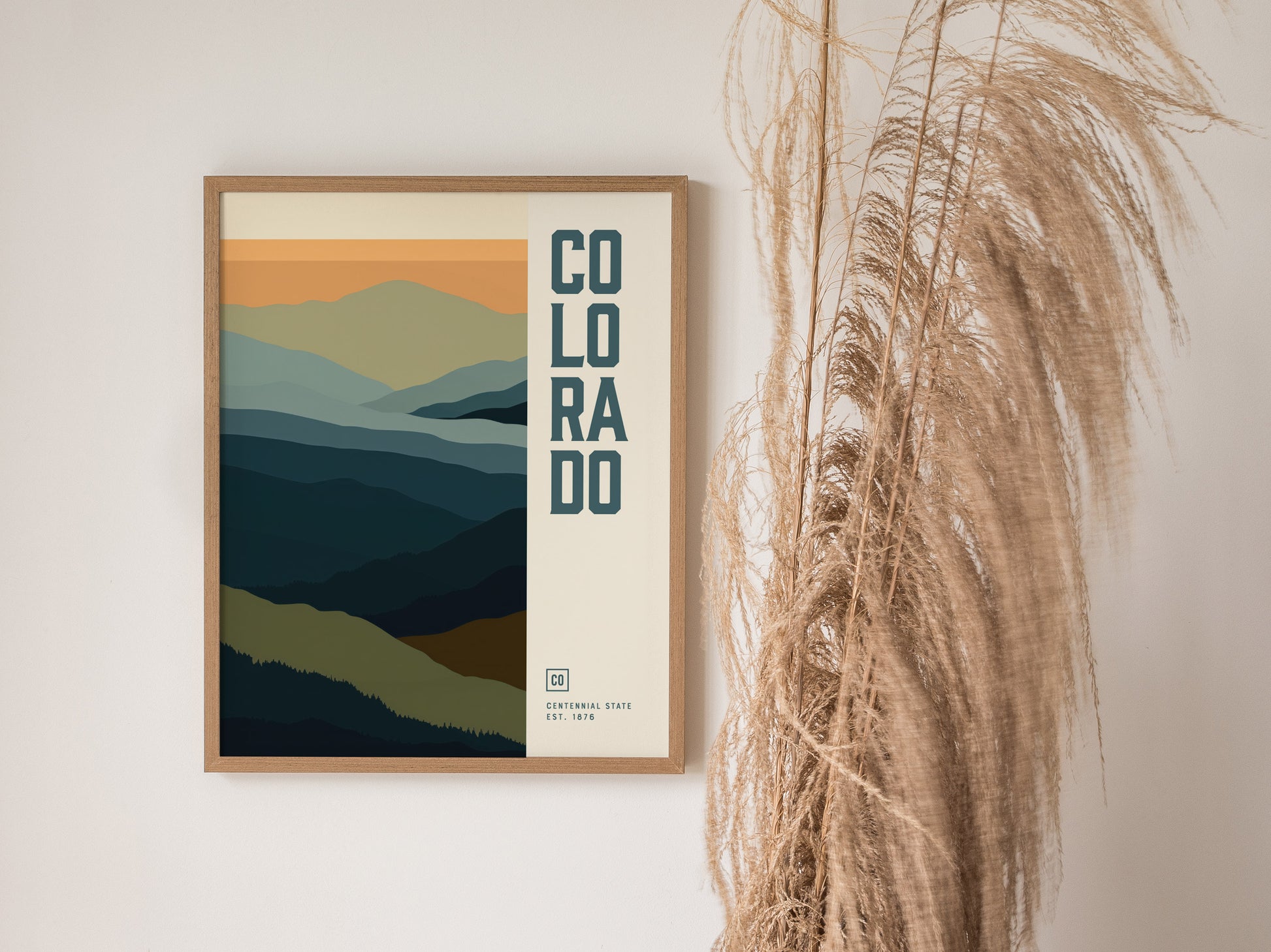 A framed poster featuring abstract layers of blue and green mountains with Colorado text is displayed next to tall, dried pampas grass against a white wall.