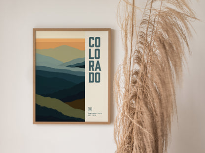 A framed poster featuring abstract layers of blue and green mountains with Colorado text is displayed next to tall, dried pampas grass against a white wall.