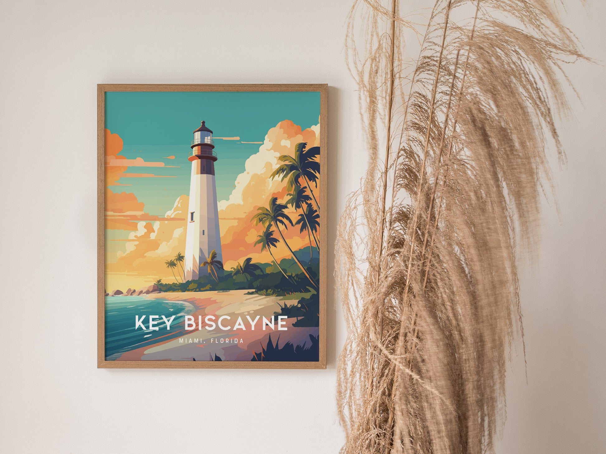 Framed Key Biscayne lighthouse poster on a wall, featuring vibrant colors of a sunset sky and swaying palm trees. Pampas grass decor is visible beside the artwork.