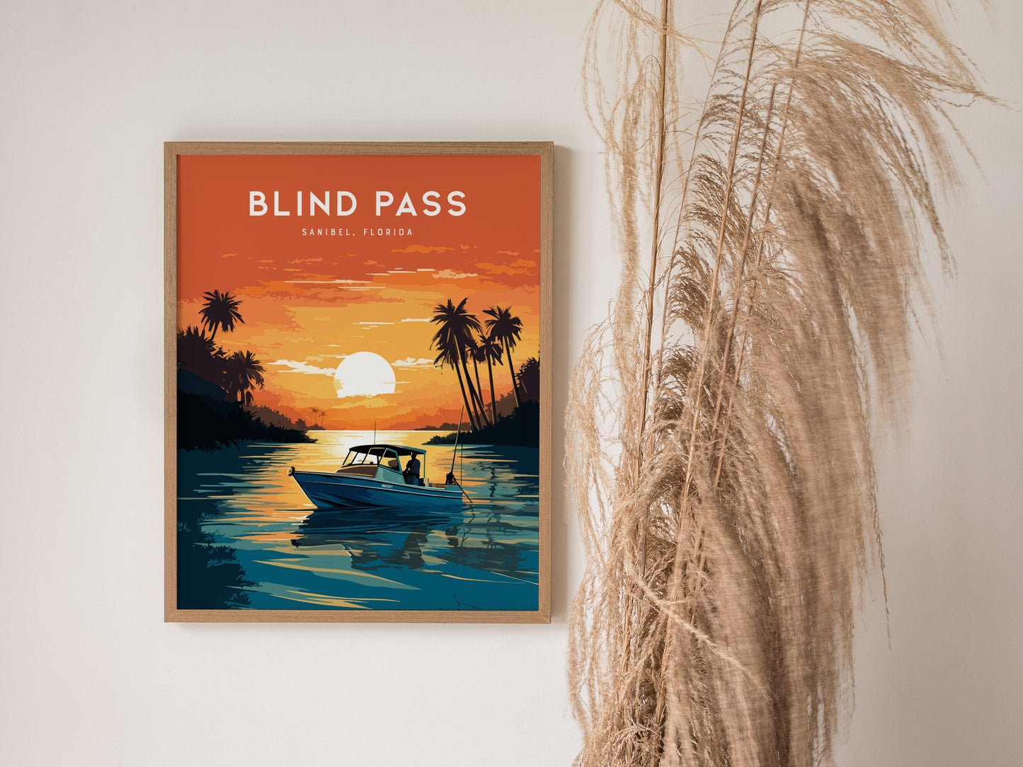 A framed poster titled Blind Pass, Sanibel, Florida depicts a boat on calm water during a vibrant orange sunset, surrounded by silhouetted palm trees. Pampas grass decorates the scene.
