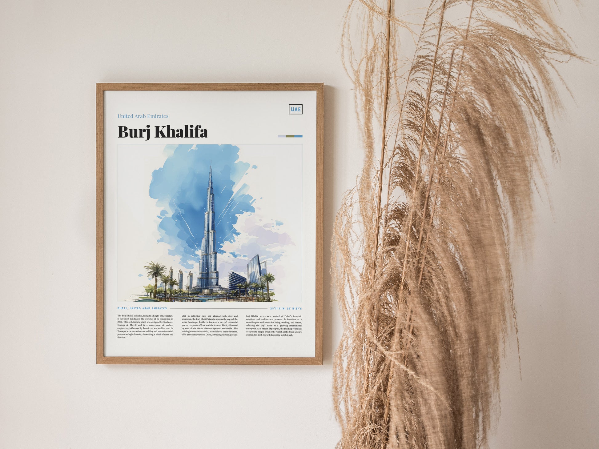 A framed poster of the Burj Khalifa with a watercolor illustration and descriptive text is displayed on a wall beside tall, dried pampas grass.