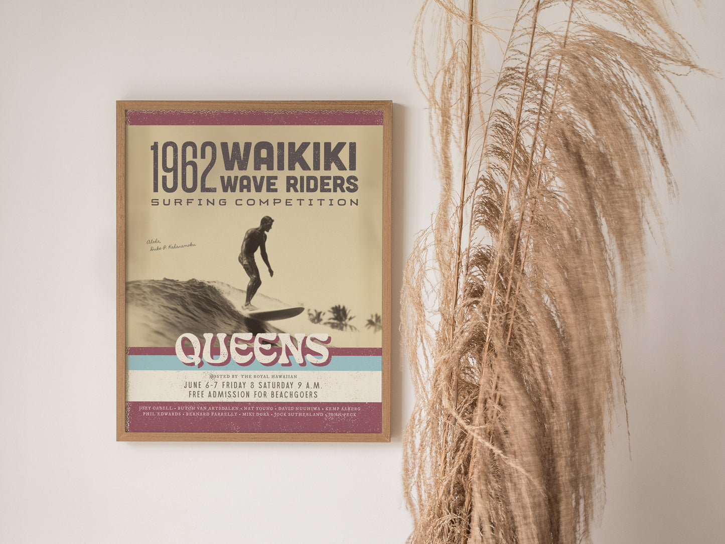 A framed vintage poster for the 1962 Waikiki Wave Riders surfing competition hangs beside dry pampas grass. The design features retro typography and a surfer riding a wave.