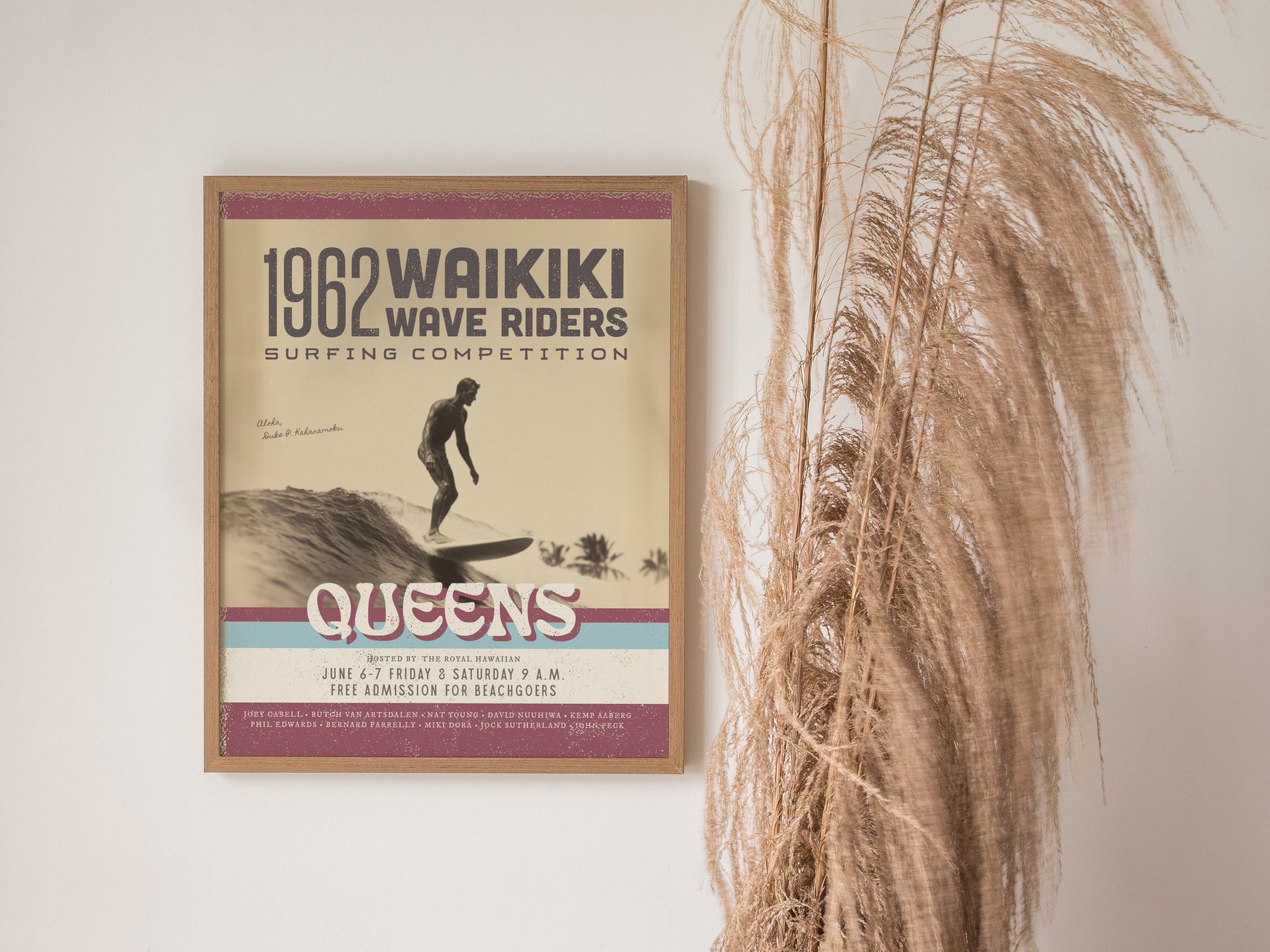 A framed vintage poster for the 1962 Waikiki Wave Riders surfing competition hangs beside dry pampas grass. The design features retro typography and a surfer riding a wave.