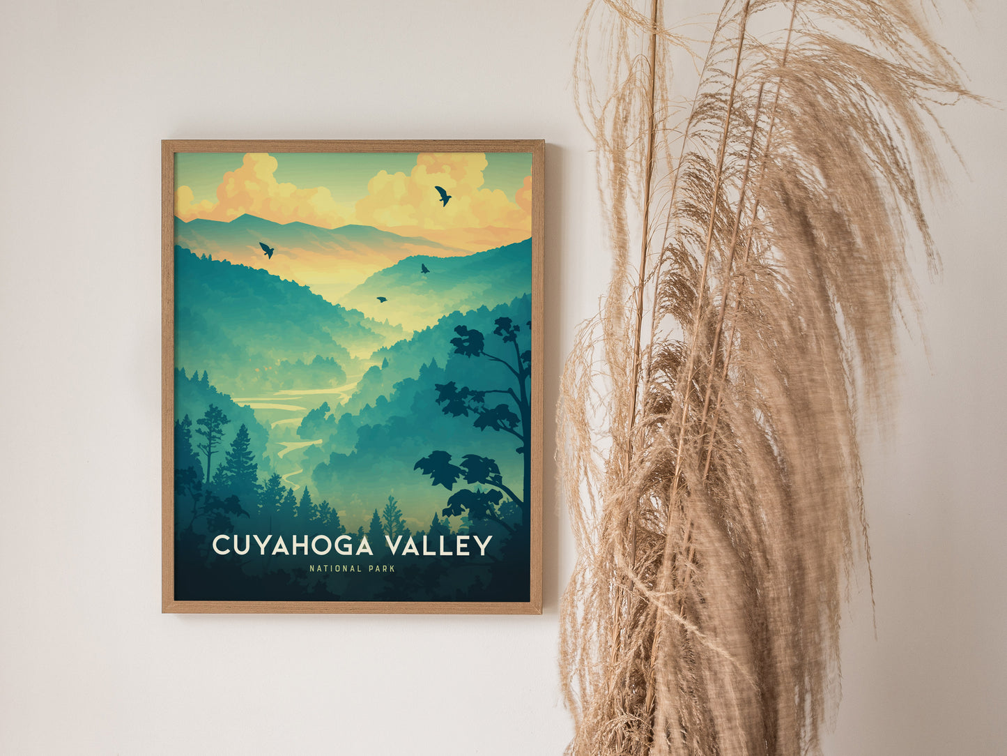 A framed poster of Cuyahoga Valley National Park features a scenic landscape with mountains, trees, and birds against a sunset sky, displayed beside dried pampas grass.