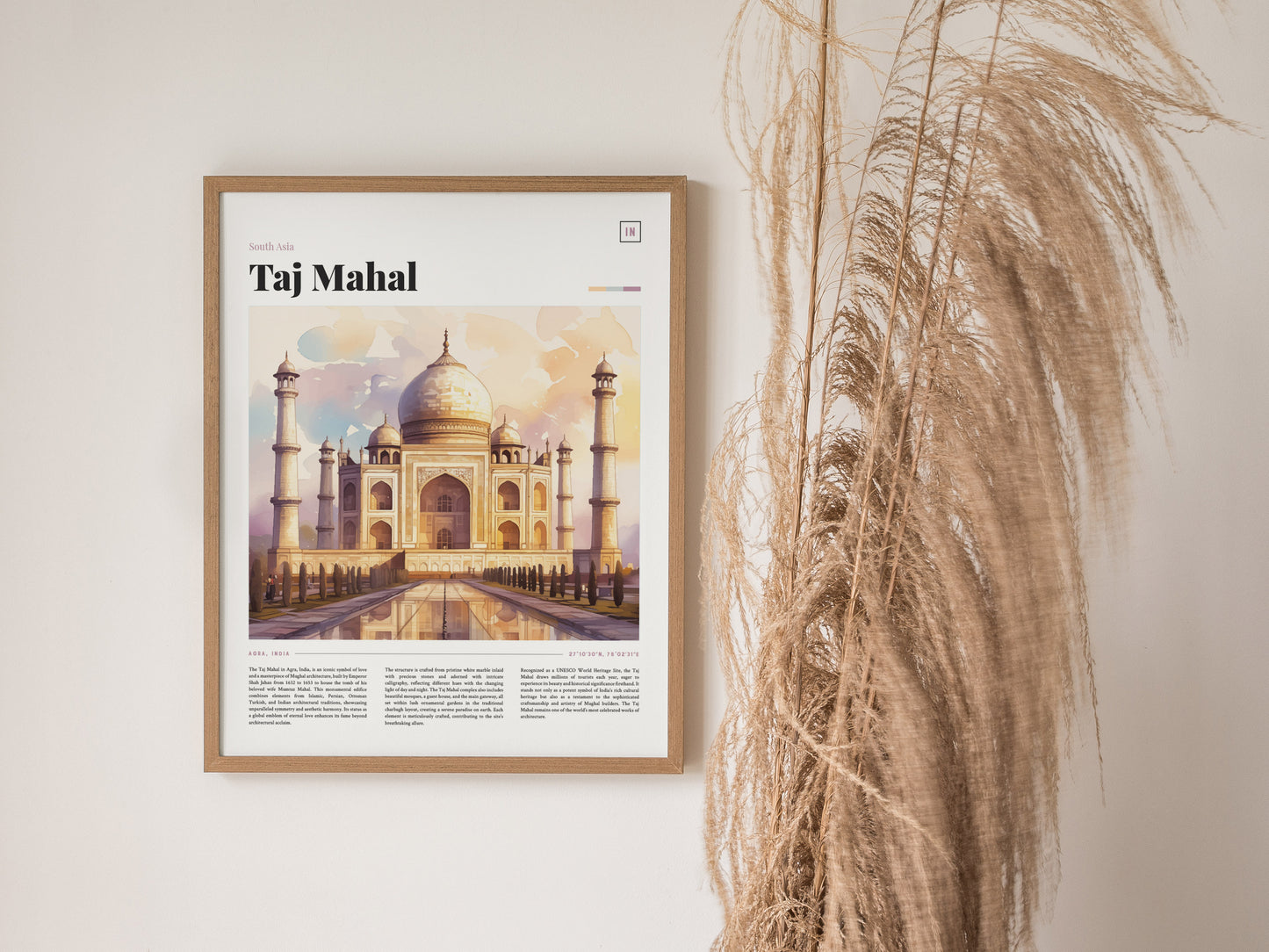 A framed poster of the Taj Mahal featuring a watercolor illustration hangs on a wall next to tall, dried grasses. The artwork highlights the monuments iconic architecture and symmetry.