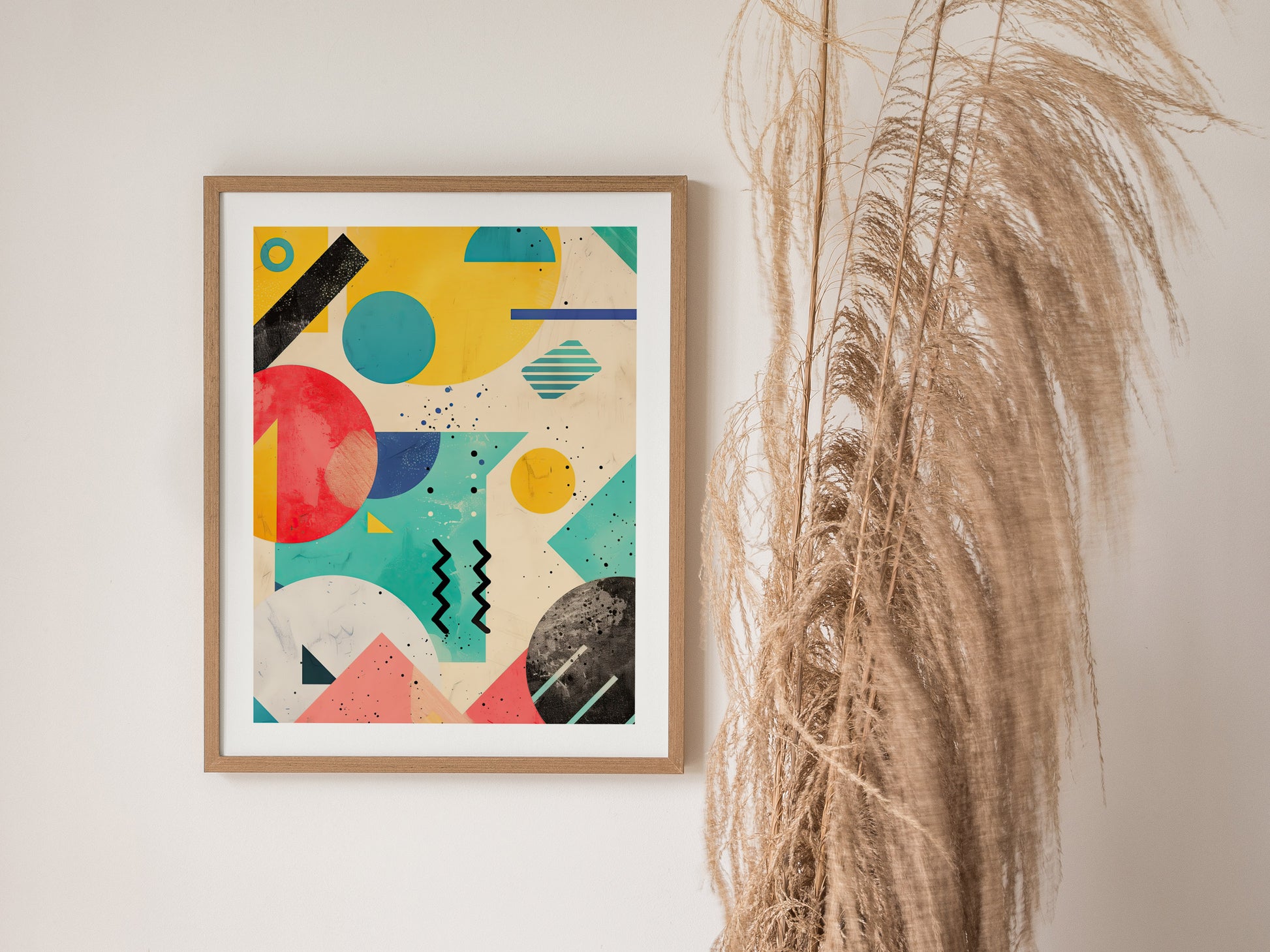A framed abstract poster featuring bold geometric shapes in vibrant colors hangs on a wall beside dried pampas grass. The artwork showcases 90s-inspired retrowave design elements.