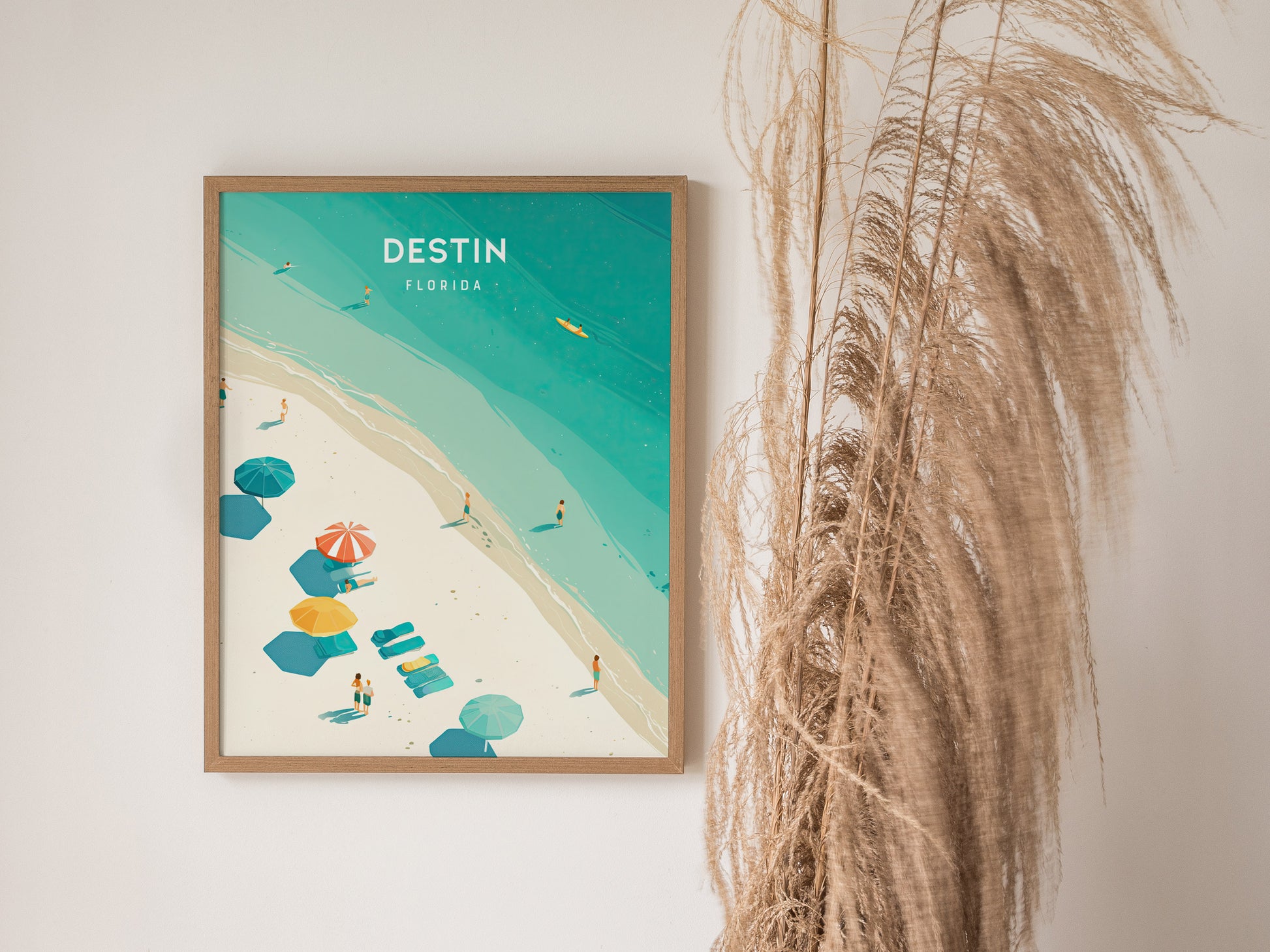 A framed poster of Destin, Florida, displaying a beach scene with turquoise waters, colorful umbrellas, and beachgoers. Dried pampas grass decorates the adjacent wall space.
