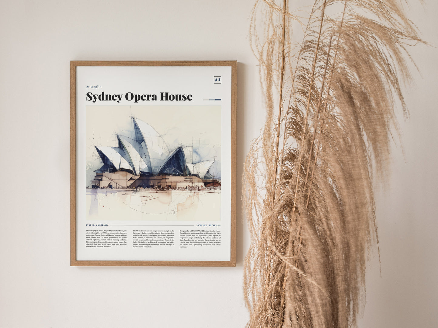 A framed poster of the Sydney Opera House hangs on a wall beside tall, dried pampas grass, showcasing a watercolor illustration of the iconic landmark.