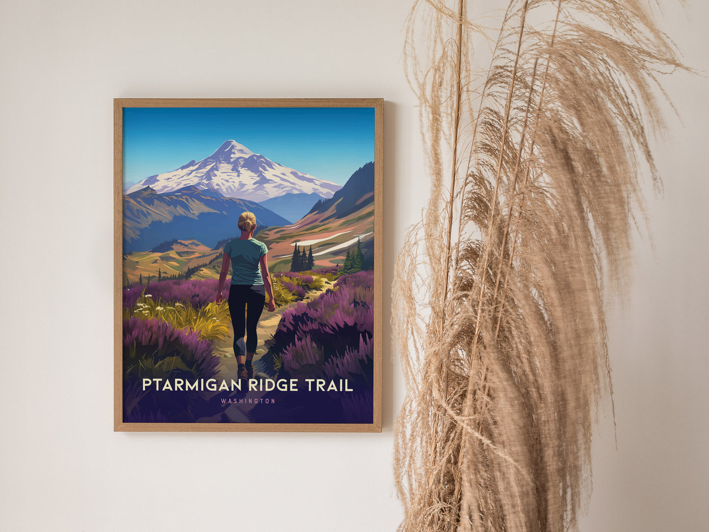 A framed poster of the Ptarmigan Ridge Trail shows a hiker in a vibrant landscape with Mt. Baker in the background. Tall dried grasses are positioned beside the poster.