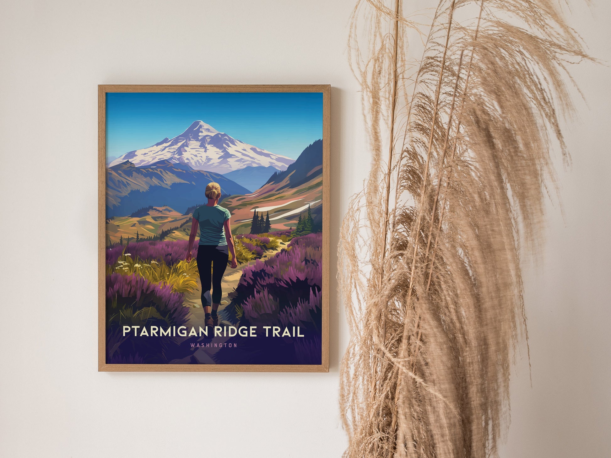 A framed poster of the Ptarmigan Ridge Trail shows a hiker in a vibrant landscape with Mt. Baker in the background. Tall dried grasses are positioned beside the poster.