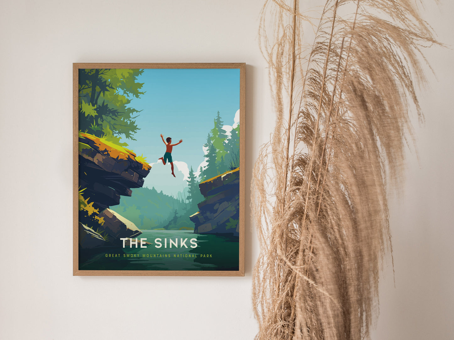 A framed poster titled The Sinks depicts a person jumping between rocky cliffs over a lush forested landscape, set against a bright blue sky. Dried pampas grass decorates the wall beside the artwork.