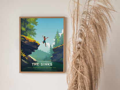 A framed poster titled The Sinks depicts a person jumping between rocky cliffs over a lush forested landscape, set against a bright blue sky. Dried pampas grass decorates the wall beside the artwork.