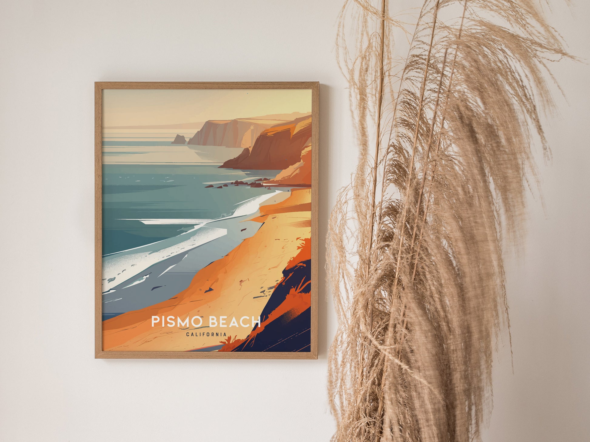 A framed poster of Pismo Beach, California, features vibrant orange cliffs and a serene ocean view. Tall dried grasses are artfully arranged beside the poster, enhancing the coastal theme.