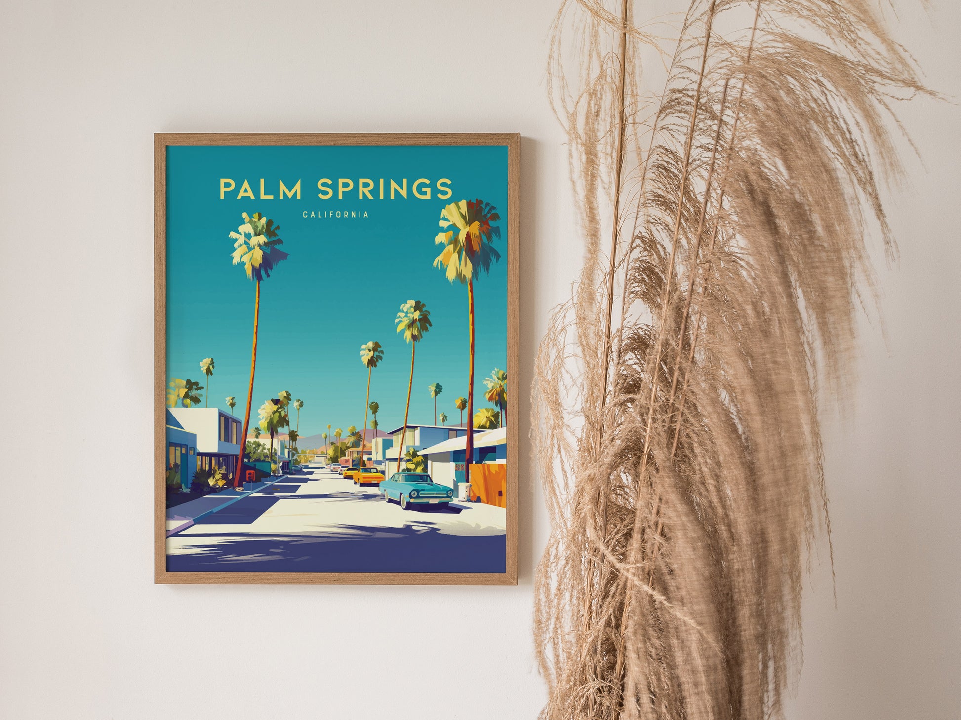 A framed Palm Springs poster features vibrant palm trees and mid-century architecture under a clear blue sky, with tall dried grasses positioned beside it for a stylish display.