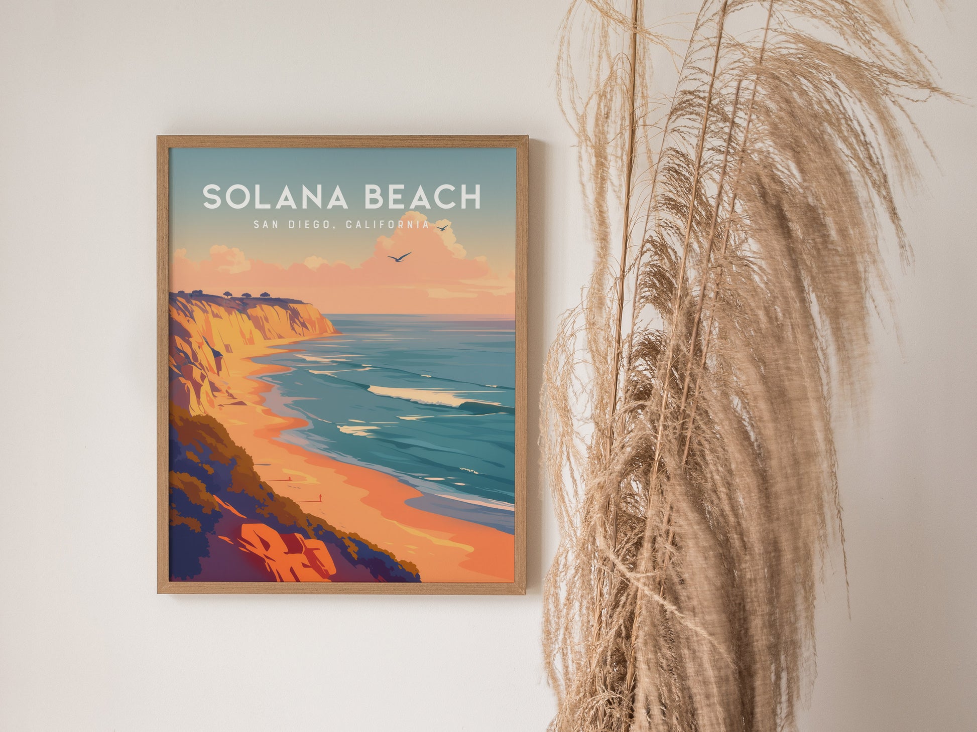 A framed Solana Beach poster features vibrant coastal bluffs, ocean waves, and a pastel sunset. Its displayed next to tall, dried decorative grasses against a white wall.