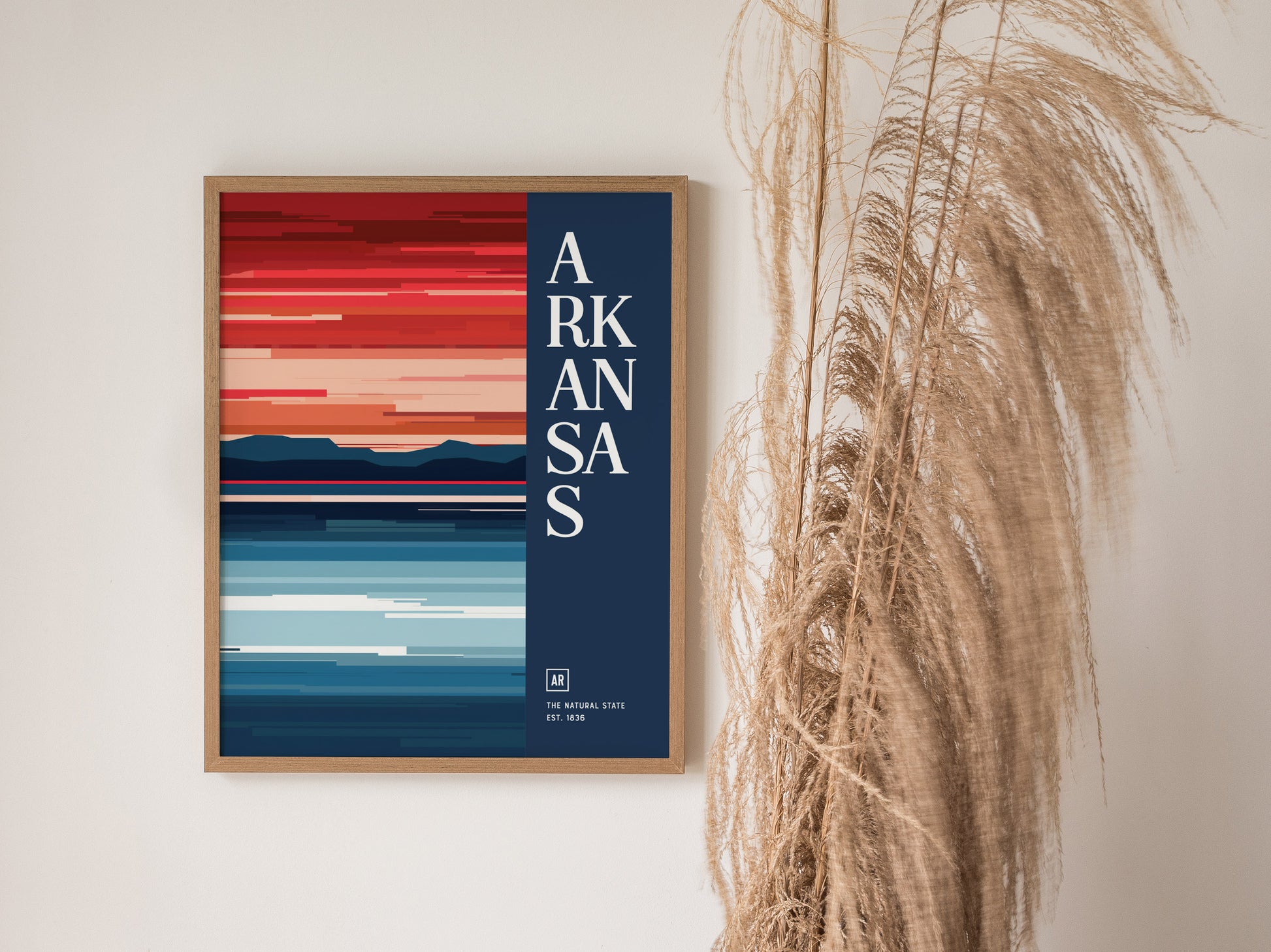 Framed Arkansas poster with bold, abstract stripes in red and blue hues, featuring Arkansas text on a navy background. Dried grasses are visible to the right of the frame.
