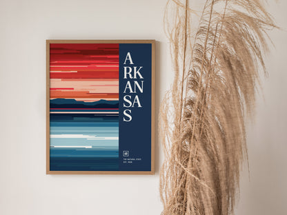 Framed Arkansas poster with bold, abstract stripes in red and blue hues, featuring Arkansas text on a navy background. Dried grasses are visible to the right of the frame.