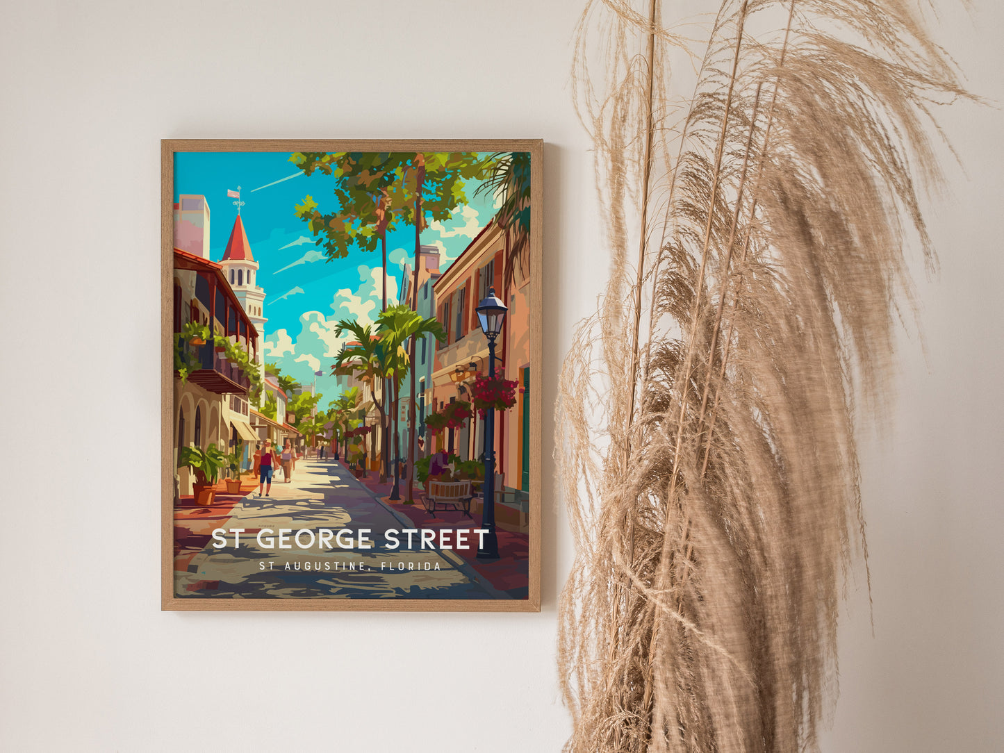 A framed illustration of St. George Street in St. Augustine, Florida, featuring colorful buildings, trees, and people walking, next to tall pampas grass against a light wall.