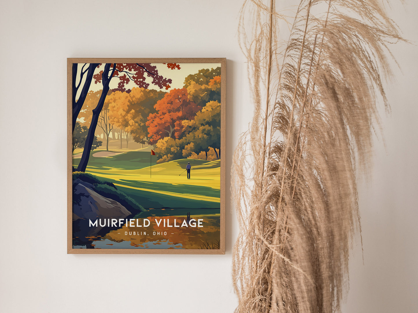 Framed artwork of Muirfield Village Golf Club with vibrant autumn trees and a golfer on the course, displayed on a wall next to tall, wispy pampas grass.