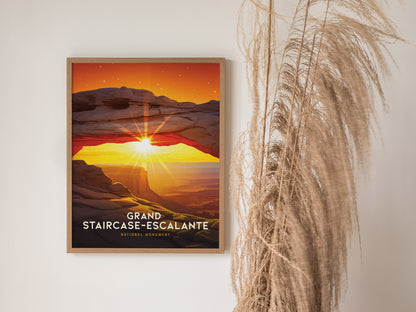 A framed poster of Grand Staircase–Escalante depicts a vibrant sunset arch with radiant sunlight. It hangs on a white wall next to tall dried ornamental grass.