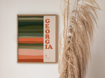 Framed Georgia poster with horizontal earthy stripes and bold typography, displayed next to decorative dried grass. The design is minimalist, featuring colors like green, orange, and peach.