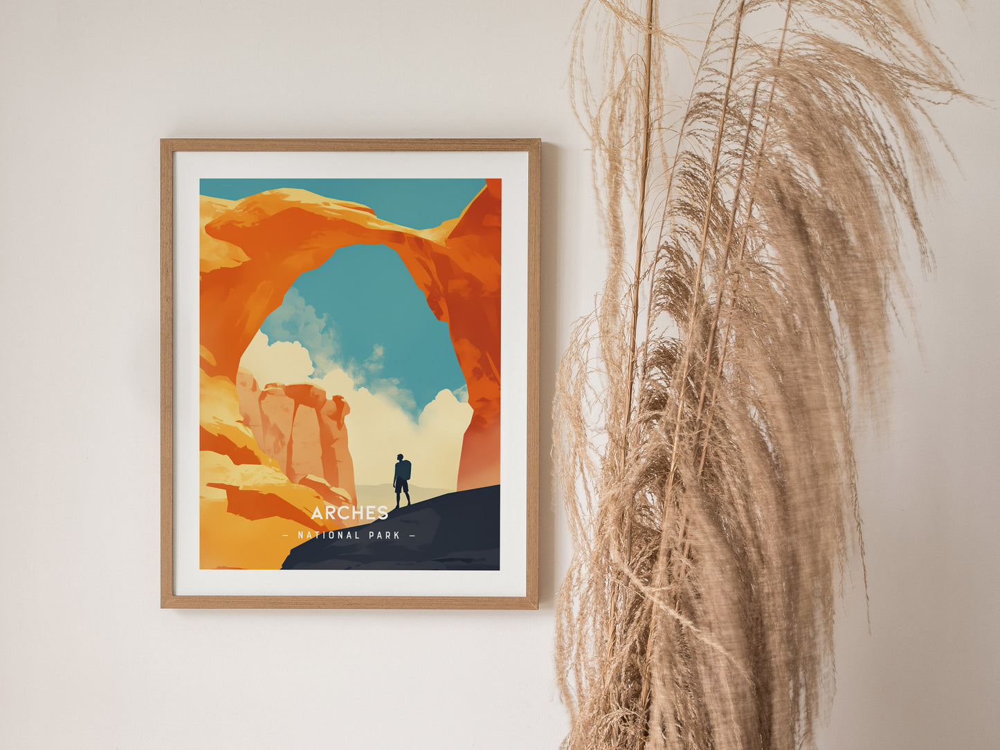 A framed minimalist poster of Arches National Park displays Delicate Arch with a lone figure beneath a vibrant sky. Tall, dried grasses are visible beside the framed artwork.