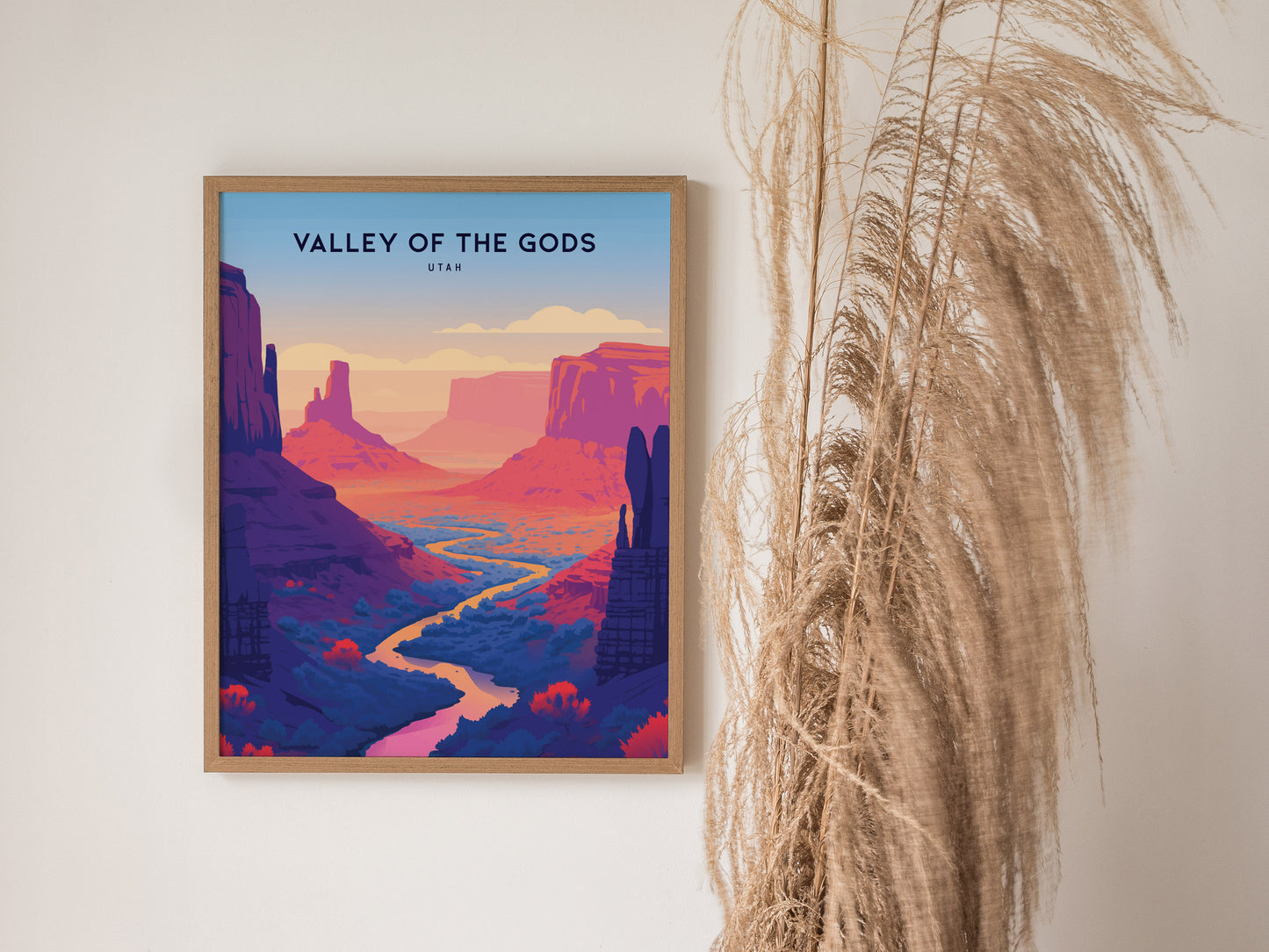 A framed Valley of the Gods poster features vibrant red and purple rock formations with a winding river, set against a minimalist background. Tall dried grasses are beside the artwork.