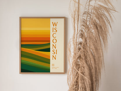 A framed Wisconsin poster with abstract layers in green, gold, and orange hangs on a white wall beside tall, dried grasses. The design features vertical text reading Wisconsin.