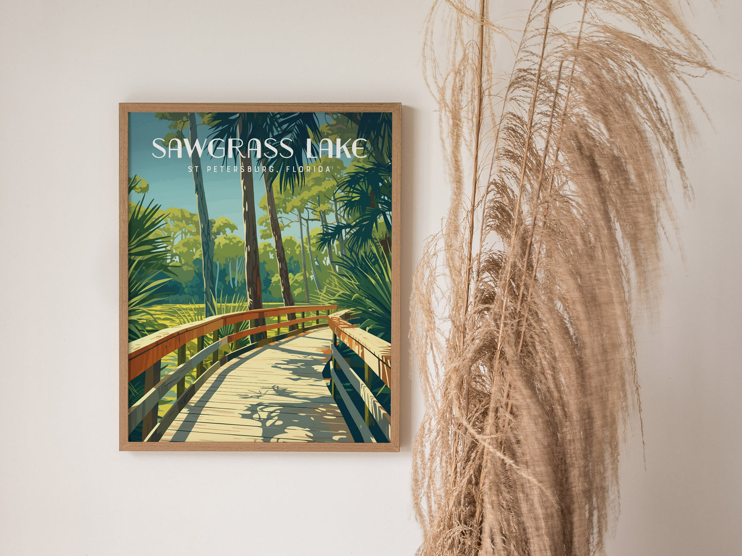 Framed poster of Sawgrass Lake Park with a wooden boardwalk surrounded by lush greenery. Decorative dried grasses are placed beside the poster against a plain white wall.