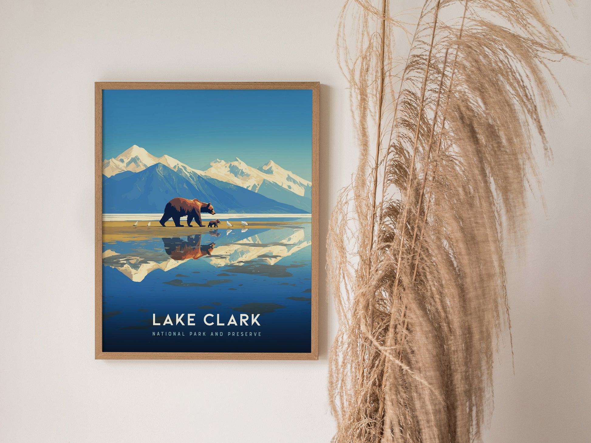 Framed artwork of Lake Clark National Park featuring a bear and cubs walking along the shore, with snow-capped mountains in the background, displayed next to tall dried grasses.
