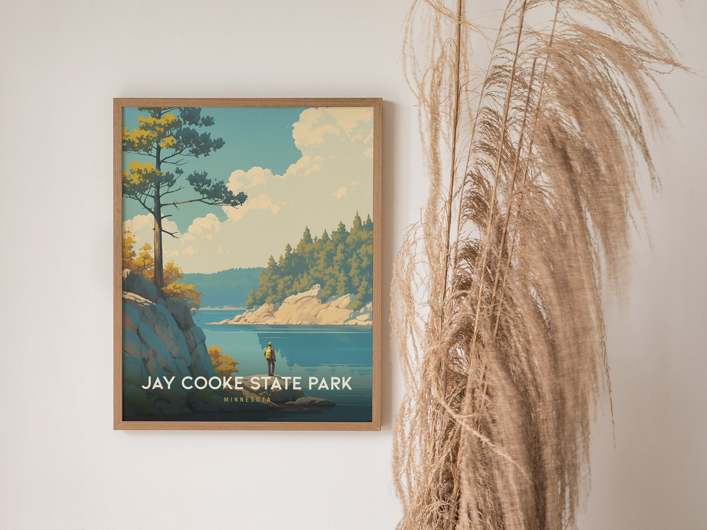 A framed Jay Cooke State Park poster hangs on a white wall, depicting a scenic landscape with trees, water, and clouds. Tall decorative grass stands beside the poster.