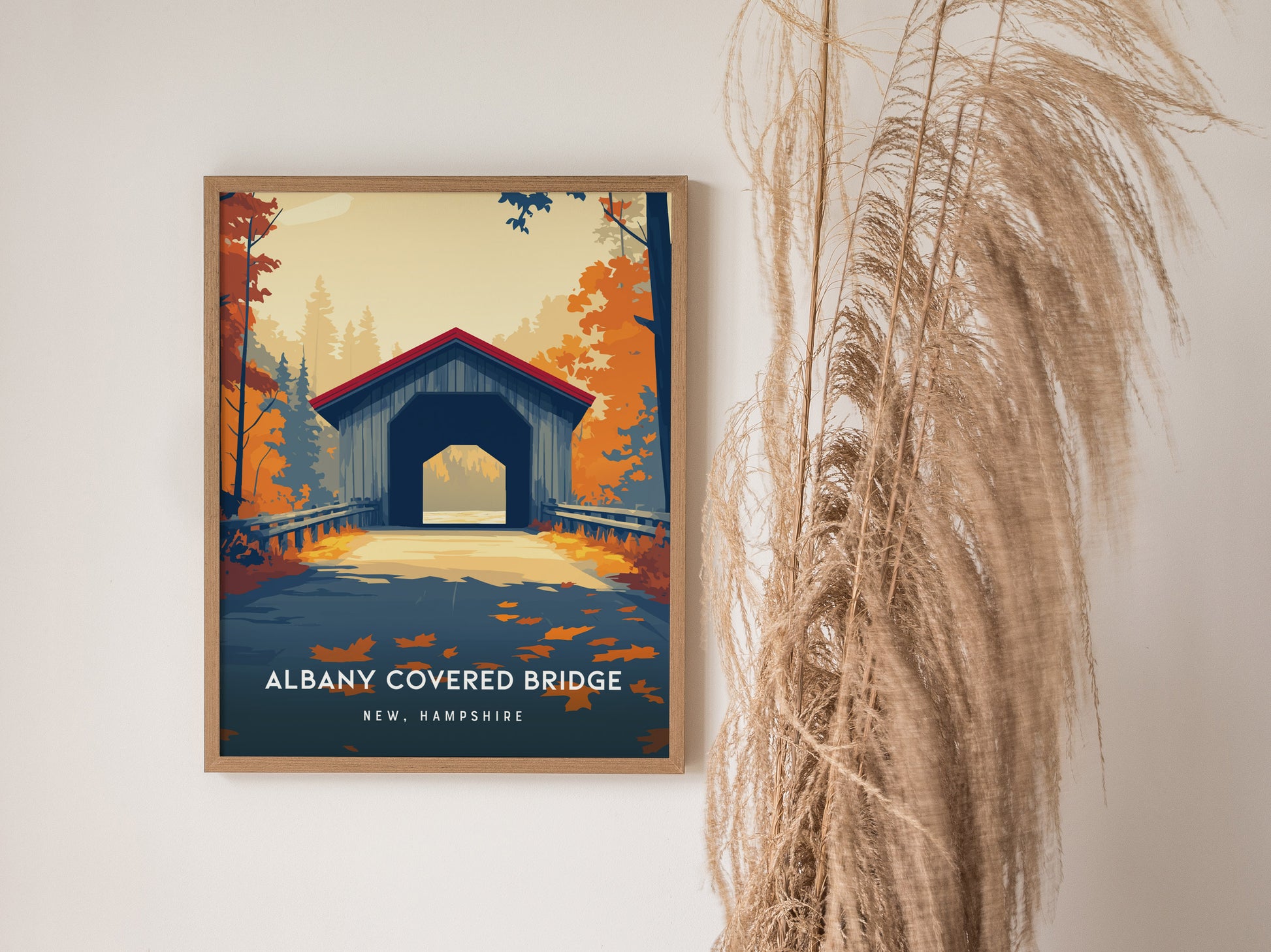A framed poster of the Albany Covered Bridge in New Hampshire is displayed against a light wall, showcasing a vibrant illustration of the bridge surrounded by colorful autumn foliage.