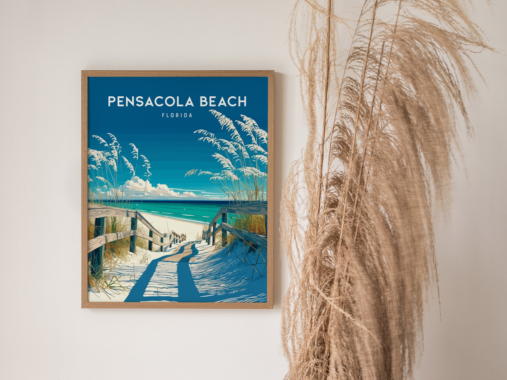 A framed Pensacola Beach poster depicting a sandy path leading to the sea, surrounded by tall grasses, hangs on a white wall beside decorative dried plants.