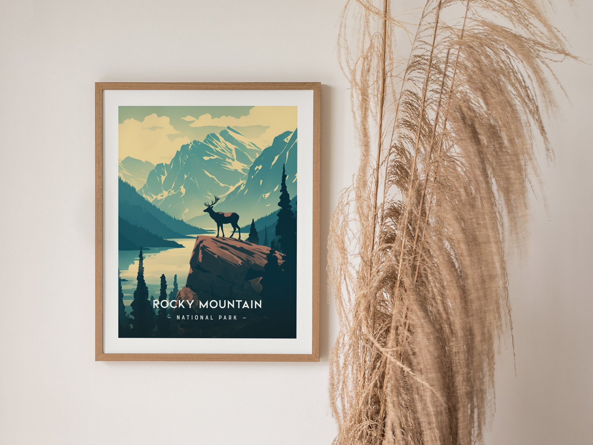 Framed Rocky Mountain National Park poster depicting a lone elk on a rock, surrounded by alpine lakes and mountains, with tall dried grasses beside the artwork.