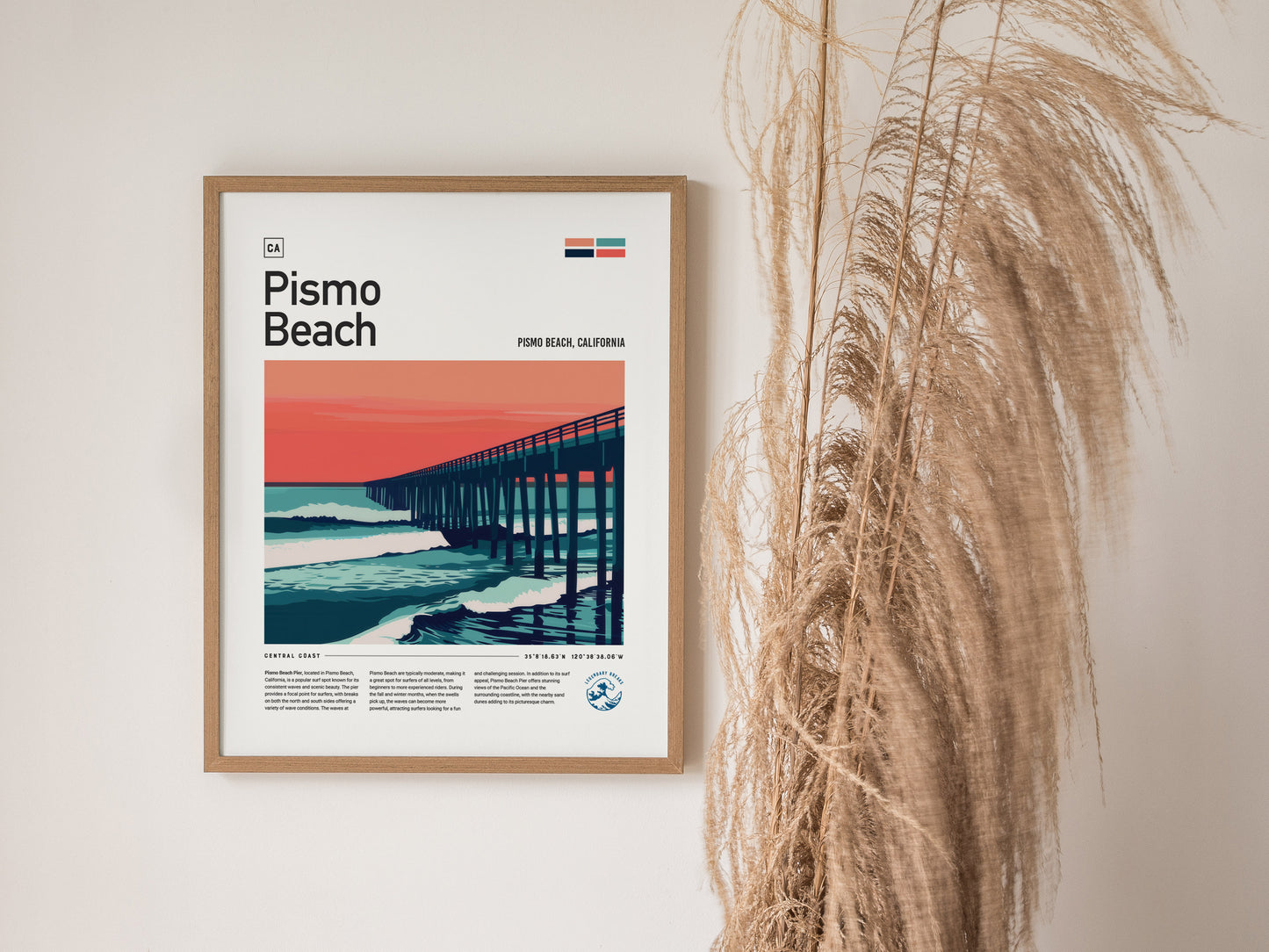 Framed Pismo Beach poster featuring a stylized pier against a vibrant sunset with ocean waves. The poster is displayed on a wall beside dried pampas grass.