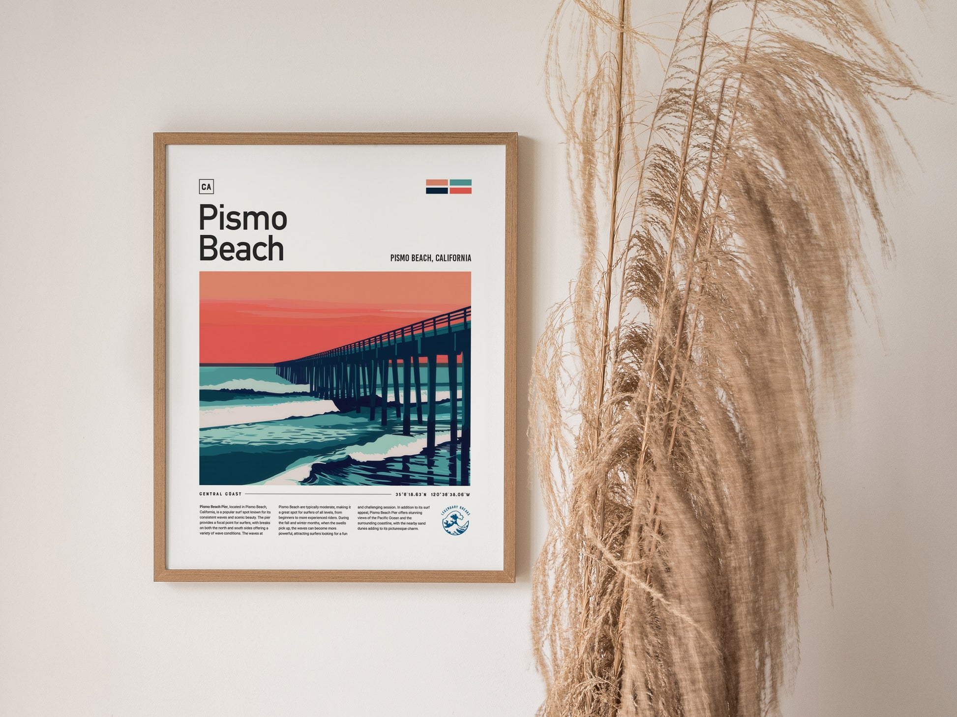 Framed Pismo Beach poster featuring a stylized pier against a vibrant sunset with ocean waves. The poster is displayed on a wall beside dried pampas grass.