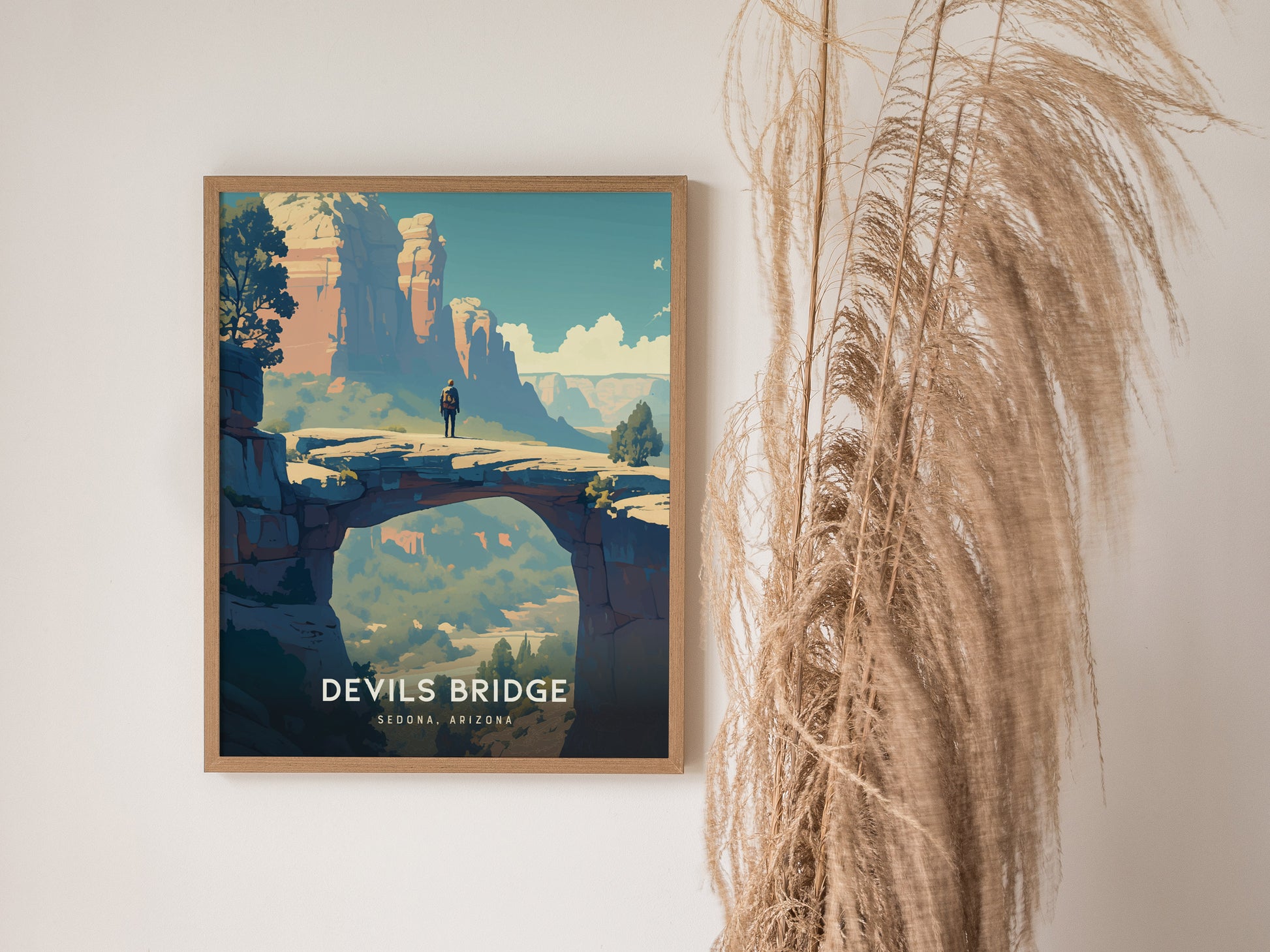 A framed poster of a person standing on Devils Bridge in Sedona, Arizona, featuring vivid desert colors and rugged cliffs, hangs on a wall beside dried pampas grass.